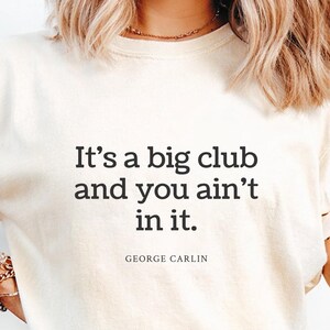 May include: Cream-colored t-shirt with the quote "It's a big club and you ain't in it" by George Carlin in black text. The shirt is worn with blue denim shorts.