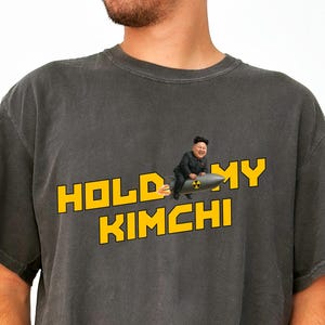 May include: Dark gray t-shirt with the text "HOLD MY KIMCHI" in yellow block letters. The graphic features a cartoon image of a person riding a missile. The shirt is a casual, unisex style.