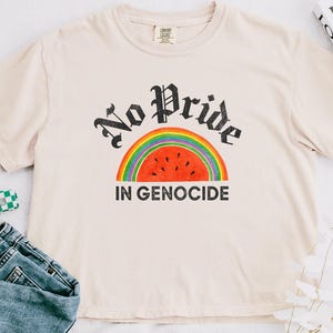 May include: A cream-coloured t-shirt featuring the text "No Pride In Genocide" in a black gothic font above a rainbow and watermelon design. The shirt is short-sleeved with a crew neck.
