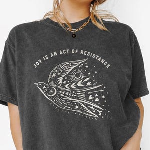 Può includere: T-shirt grigio scuro con la scritta "JOY IS AN ACT OF RESISTANCE" sopra un'illustrazione bianca dettagliata di un uccello con elementi celesti. Il design dell'uccello include stelle, un sole e altri dettagli decorativi.