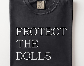May include: A black short-sleeved t-shirt featuring the white text "PROTECT THE DOLLS" printed in a serif font on the front. The shirt has a crew neckline and a Comfort Colors brand label visible on the inside of the collar, indicating a size medium.