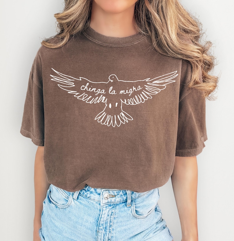 May include: Brown t-shirt with a white graphic of a dove with outstretched wings and the text "chinga la migra". The shirt has short sleeves and a crew neck. The model is wearing blue denim shorts.