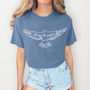May include: A blue t-shirt featuring a white graphic of a dove with outstretched wings. The text "chinga la migra" is written in cursive within the dove's wings. The shirt is paired with denim shorts.