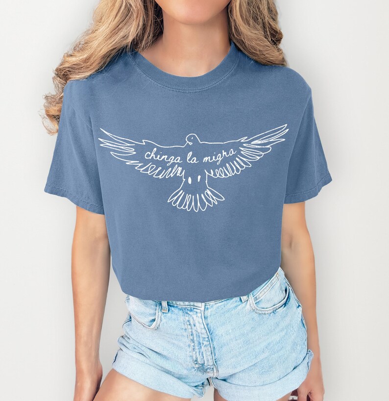 May include: A blue t-shirt featuring a white graphic of a dove with outstretched wings. The text "chinga la migra" is written in cursive within the dove's wings. The shirt is paired with denim shorts.