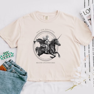Puede incluir: Camiseta color crema con una ilustración en blanco y negro de una niña montando un unicornio. El texto "MAY OUR DAUGHTERS BECOME UNGOVERNABLE" está impreso encima y debajo de la imagen. La camiseta es de material suave.