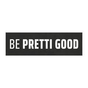 May include: A rectangular black sign featuring the phrase "BE PRETTI GOOD" in bold, white capital letters. The sign has a minimalist design, with the text centrally positioned against the dark backdrop.