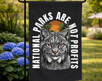 May include: Black garden flag with the text "NATIONAL PARKS ARE NOT PROFITS" in white. The flag features a detailed illustration of a bobcat's face, with orange eyes, set against a backdrop of a sunset and palm trees.