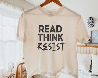 May include: A cream-colored t-shirt with the words "READ THINK RESIST" printed in black block letters. The shirt is hanging on a wooden hanger. The text is centered on the front of the shirt.