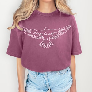 May include: A mauve-colored t-shirt with a white graphic of a dove with outstretched wings. The text "chinga la migra" is written across the dove's wings. The shirt has short sleeves and a crew neck.