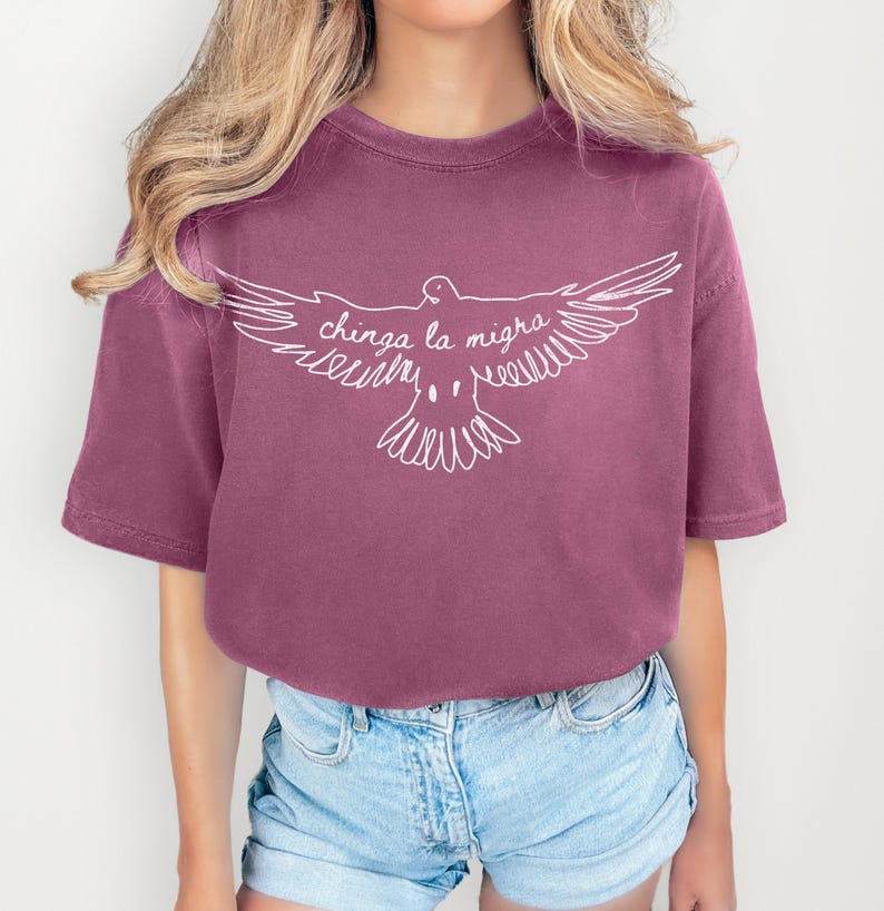 May include: A mauve-colored t-shirt with a white graphic of a dove with outstretched wings. The text "chinga la migra" is written across the dove's wings. The shirt has short sleeves and a crew neck.
