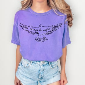 May include: A lilac-colored t-shirt featuring a black line drawing of a dove with outstretched wings. The text "chinga la migra" is written above the dove. The shirt is paired with light blue denim shorts.
