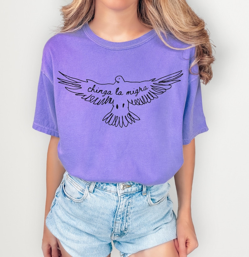 May include: A lilac-colored t-shirt featuring a black line drawing of a dove with outstretched wings. The text "chinga la migra" is written above the dove. The shirt is paired with light blue denim shorts.