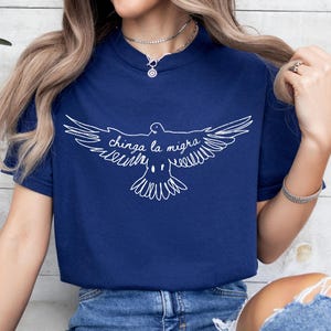 May include: Navy blue t-shirt featuring a white graphic of a dove with outstretched wings and the text "chinga la migra". The design is centered on the front of the shirt. The shirt has a crew neck and short sleeves.