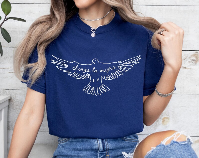 May include: Navy blue t-shirt featuring a white graphic of a dove with outstretched wings and the text "chinga la migra". The design is centered on the front of the shirt. The shirt has a crew neck and short sleeves.