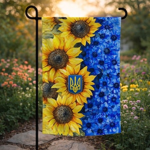 Op de afbeelding: Een tuinvaandel met een gesplitst ontwerp. De linkerkant heeft gele zonnebloemen, de rechterkant blauwe bloemen. Een blauw en geel embleem is gecentreerd op de zonnebloemen. De vlag wordt buiten getoond.