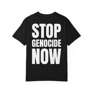 Op de afbeelding: Zwart t-shirt met de witte tekst "STOP GENOCIDE NOW" in grote letters. De tekst staat gecentreerd op de achterkant van het shirt. Het shirt heeft korte mouwen en een ronde hals.
