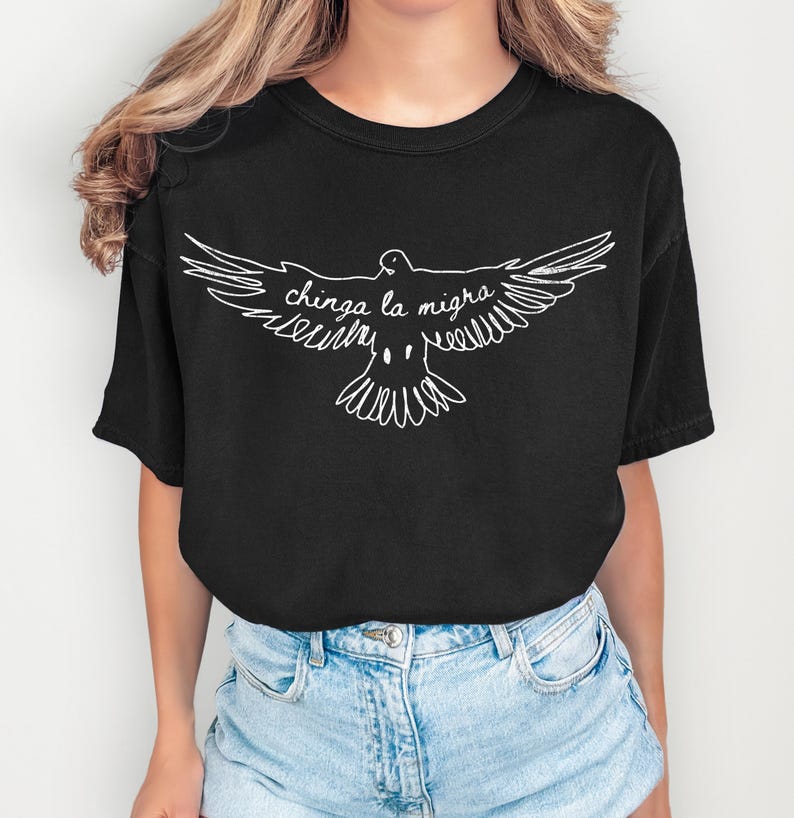May include: Black t-shirt featuring a white outline of a dove with outstretched wings. The text "chinga la migra" is written in cursive within the dove's silhouette. The shirt has short sleeves and a crew neck.