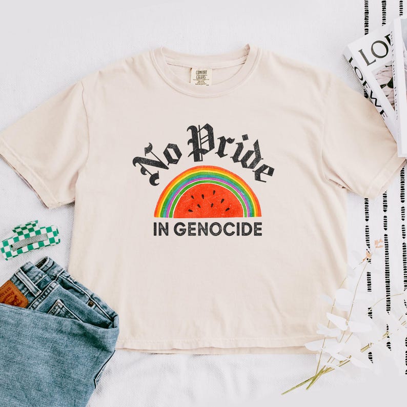 May include: A cream-coloured t-shirt featuring the text "No Pride In Genocide" in a black gothic font above a rainbow and watermelon design. The shirt is short-sleeved with a crew neck.