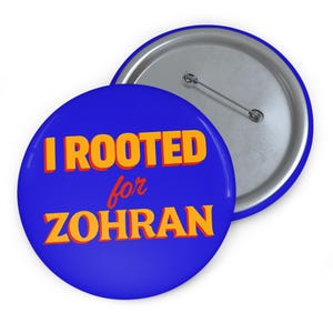May include: A blue button with the text "I ROOTED for ZOHRAN" in orange letters. The button has a silver backing with a pin. The button is round and has a blue border.
