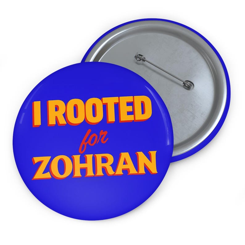 May include: A blue button with the text "I ROOTED for ZOHRAN" in orange letters. The button has a silver backing with a pin. The button is round and has a blue border.