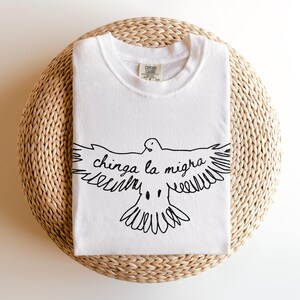 May include: White t-shirt with a black graphic of a dove with outstretched wings and the text "chinga la migra". The shirt is folded and placed on a woven, circular surface. The shirt has a crew neck and short sleeves.