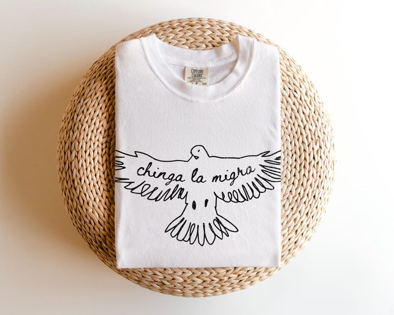 May include: White t-shirt with a black graphic of a dove with outstretched wings and the text "chinga la migra". The shirt is folded and placed on a woven, circular surface. The shirt has a crew neck and short sleeves.