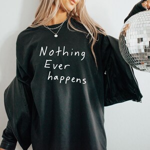 May include: Black long-sleeve shirt with the white text "Nothing Ever Happens". The shirt is a dark color and has a relaxed fit. The image also shows a silver necklace and a disco ball.