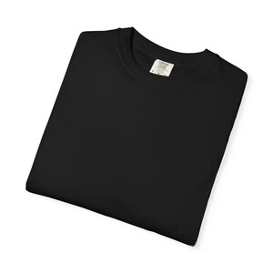 Op de afbeelding: Een gevouwen zwart t-shirt met een ronde hals. Het shirt heeft een label op de kraag met de woorden "COMFORT COLORS" erop gedrukt. Het shirt is gemaakt van een zacht materiaal en is netjes opgevouwen.