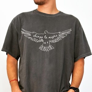 May include: Dark gray t-shirt featuring a white outline of a dove with outstretched wings. The text "chinga la migra" is written across the dove's wings. The shirt has short sleeves and a crew neck.