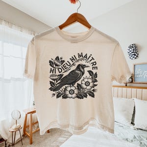 May include: Cream-colored t-shirt featuring a black and white graphic of a raven surrounded by flowers, with the text "NI DIEU NI MAITRE" in a circular design. The shirt is hanging on a wooden hanger.
