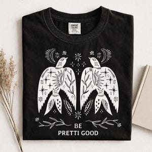 May include: Black t-shirt with a white graphic of two birds with star and moon details, and the text "BE PRETTI GOOD". The shirt has a crew neck and a Comfort Colors tag.
