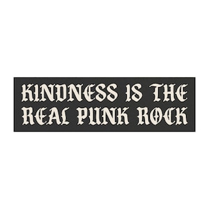 May include: A rectangular black sticker with the text "KINDNESS IS THE REAL PUNK ROCK" in a gothic font. The text is in off-white, contrasting against the dark background. The sticker is a statement piece.