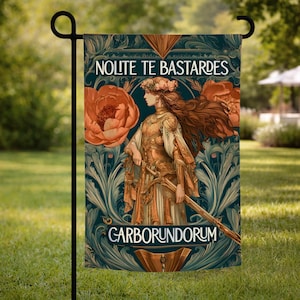 May include: A garden flag with a design of a woman in a golden gown, holding a staff, with the text "Nolite Te Bastardes Carborundorum." The design includes orange flowers and teal foliage against a dark teal background.