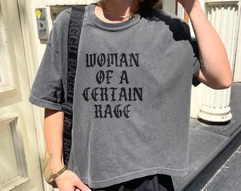 May include: A gray, short-sleeved t-shirt with the phrase "WOMAN OF A CERTAIN RAGE" printed in black gothic lettering. The shirt has a slightly faded appearance. The model is wearing a black skirt and a shoulder bag.