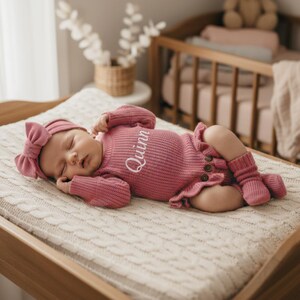 May include: A baby wearing a pink knitted outfit, including a headband with a bow, a long-sleeved onesie embroidered with the name "Quinn", and matching socks. The outfit has a ruffled skirt and wooden buttons. The baby is lying on a cream knitted blanket.