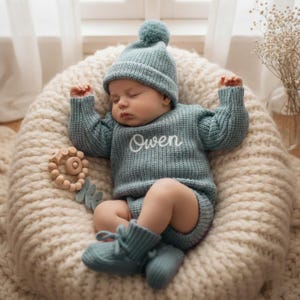 May include: A sleeping baby dressed in a teal knitted outfit, including a hat, sweater, shorts, and booties. The sweater is embroidered with the name "Owen". A wooden teething toy is placed nearby. The baby rests on a cream-coloured, knitted surface.