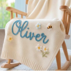 Könnte beinhalten: Eine cremefarbene Strickdecke mit dem in blauer Schreibschrift eingestickten Namen Oliver. Die Decke ist mit kleinen, dreidimensionalen gehäkelten Blumen in Weiß und Hellblau sowie einer kleinen, flauschigen Hummel-Applikation verziert.