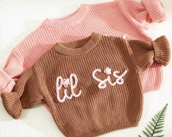 May include: Two knitted sweaters, one brown and one pink, with the words "Lil Sis" embroidered in pink on the brown sweater.