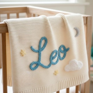 May include: Cream knitted baby blanket with the name "Leo" in blue script. The blanket has cloud and star embellishments, and is draped over a wooden cot.