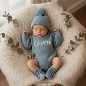 May include: A newborn baby wearing a light blue knitted outfit, including a hat with a pom-pom, a long-sleeved onesie embroidered with the name "Owen", and matching booties. The baby is lying on a cream-coloured knitted surface.