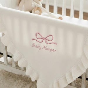 Può includere: Coperta per neonati color crema con un fiocco rosa e la scritta ricamata "Baby Harper". La coperta ha un bordo a balze ed è drappeggiata su una culla. Un peluche è visibile sullo sfondo.
