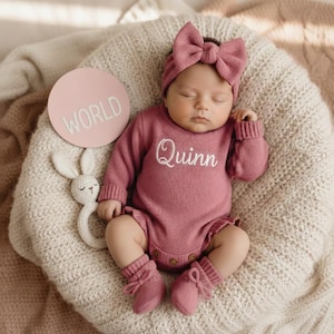 May include: A sleeping baby in a dusty rose knitted outfit, featuring a onesie embroidered with "Quinn", a bow headband, and matching socks. A round sign with "WORLD" and a white bunny rattle are also present. The outfit is made of soft knit fabric.