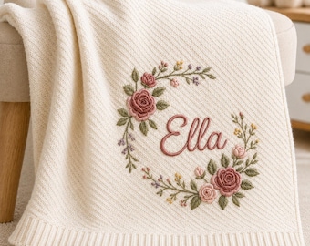 May include: A cream-colored knit blanket featuring a diagonal ribbed texture and a wide ribbed border. The center displays an embroidered floral wreath in shades of pink, green, and yellow, encircling the name Ella stitched in pink script.