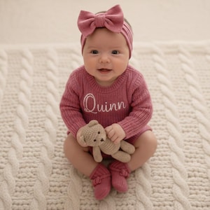 May include: A baby in a pink knitted outfit, featuring a sweater embroidered with the name "Quinn", a bow headband, and matching socks. The baby holds a small teddy bear, sitting on a white cable-knit blanket. The outfit is a soft, warm colour.