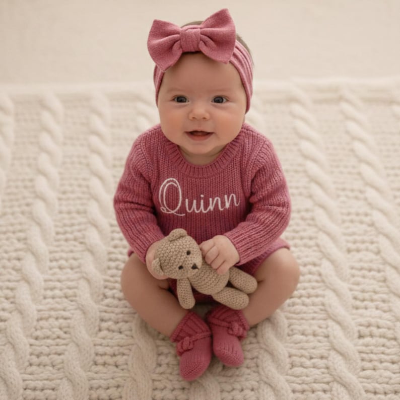 May include: A baby in a pink knitted outfit, featuring a sweater embroidered with the name "Quinn", a bow headband, and matching socks. The baby holds a small teddy bear, sitting on a white cable-knit blanket. The outfit is a soft, warm colour.