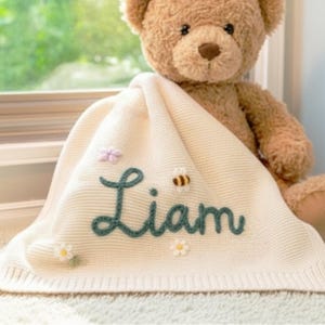 Op de afbeelding: Crèmekleurige babydeken met de naam "Liam" geborduurd in teal schrift. De deken is versierd met een kleine bij, bloemen en een vlinder. Een teddybeer is gedeeltelijk zichtbaar op de achtergrond.