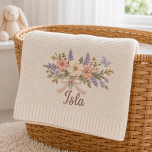 Può includere: Una coperta in maglia color crema con un bouquet ricamato di fiori rosa, bianchi e color lavanda con foglie verdi e una piccola farfalla. Sotto la composizione floreale, il nome Isla è ricamato in un carattere corsivo marrone.