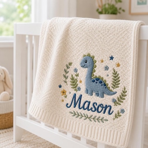 Pode incluir: Uma manta de malha cor de creme com um design bordado de um dinossauro azul rodeado por folhagem verde, pequenas flores e estrelas. O nome Mason está bordado em letra cursiva azul-escura abaixo do dinossauro, emoldurado por ramos.