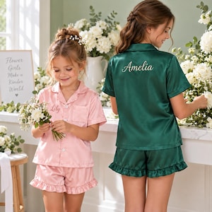 May include: A light pink satin pajama set with the name Lily embroidered on the pocket and a dark green satin pajama set with the name Amelia embroidered on the back. Both sets feature short sleeves, button-down tops, and ruffled shorts.