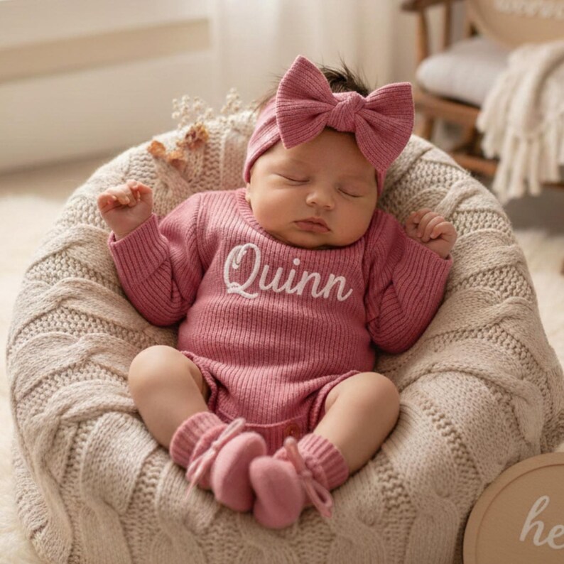 May include: A sleeping baby in a pink knitted outfit, comprising a romper, headband, and booties. The romper features the name "Quinn" embroidered on the front. The baby rests in a soft, textured, light-coloured basket.